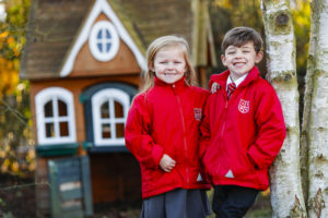A male and female pupil are shown standing with one another for a photo outside the school building. Both pupils are wearing red High Halstow jackets.