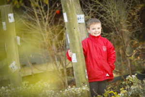 A young boy is seen standing by a tree on the school grounds, dressed in a red High Halstow jacket and smiling for the camera.