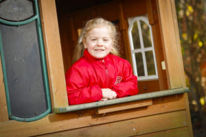 A young girl is seen dressed in a red jacket and smiling, looking out at the camera from a wooden shed window.