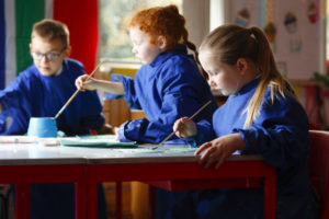 Three young pupils are pictured painting in class together, wearing Navy blue aprons.