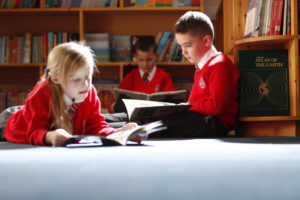 Three young pupils are seen reading books in the school library area.